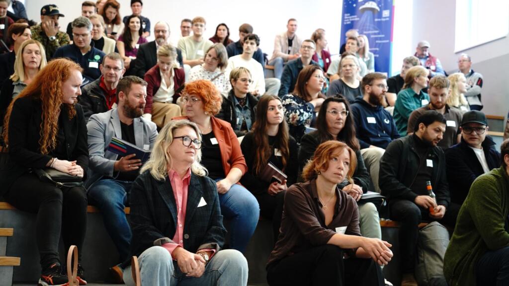 Viele Menschen sitzen in einem Atrium und schauen nach vorne.