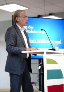 Ein Mann steht am Rednerpult. Im Hintergrund steht ein großer Bildschirm.